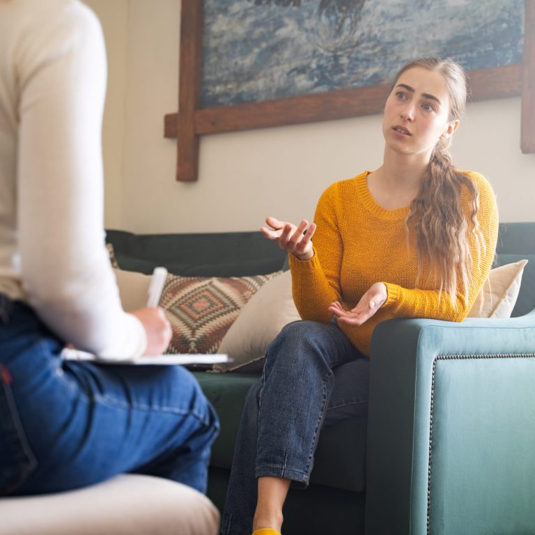 Young woman talking to psychologist during therapy session, sitting on sofa, female sharing problems while psychologist listening to her, mental health support, counseling in cozy modern interior